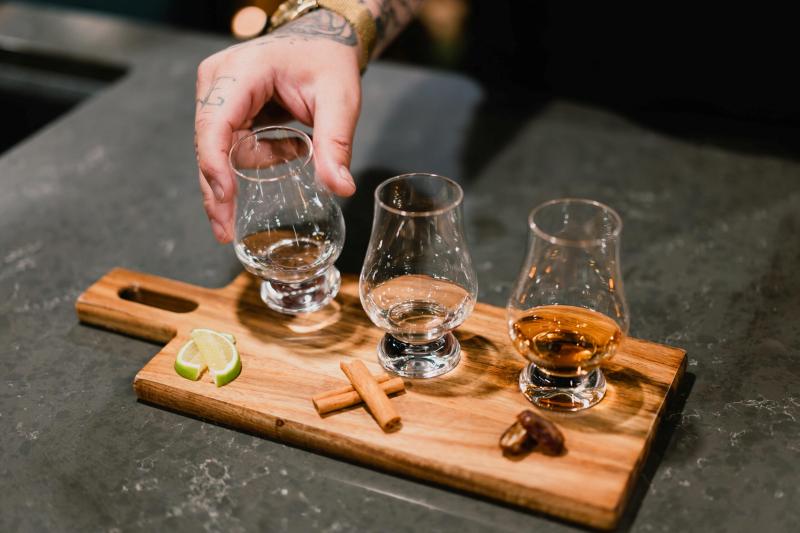 A hand with tattoos holds a whiskey glass on a tray with cinnamon sticks and lime.